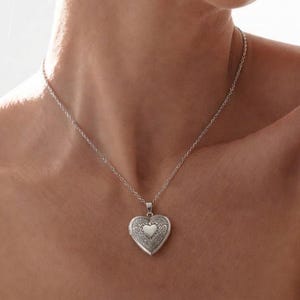 Op de afbeelding: Een zilverkleurige hartvormige medaillonhanger aan een fijne bijpassende ketting. Het medaillon is voorzien van een ingewikkeld gegraveerd patroon over het oppervlak, met in het midden een glad, gepolijst hartvormig detail.