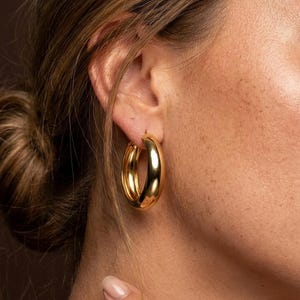 Puede incluir: Un pendiente de aro grueso de oro pulido con una superficie lisa y redondeada. El pendiente presenta un acabado metálico de alto brillo y un diseño circular que cuelga del lóbulo de la oreja, reflejando la luz en su superficie curva.