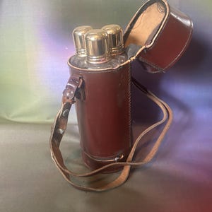 Peut inclure: Un étui de transport en cuir marron contenant trois bouteilles en verre avec des bouchons en métal doré brillant. L'étui cylindrique est doté d'un couvercle à charnière et d'une bandoulière réglable, les bouteilles étant visibles par le haut.