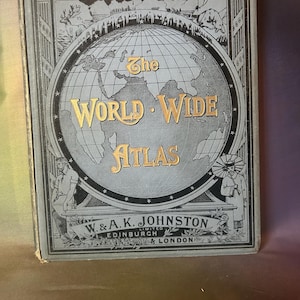 May include: A vintage hardcover book titled The World Wide Atlas by W. & A.K. Johnston, Edinburgh & London. The grey cover features a central globe illustration with gold lettering, flanked by figures and decorative borders in black and grey.