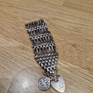 Puede incluir: Una pulsera de metal plateado con un diseño de eslabones tejidos de múltiples hilos. La pulsera incluye dos dijes colgantes: un dije circular con un patrón de cruz en relieve y un dije en forma de corazón con una superficie lisa y pulida.