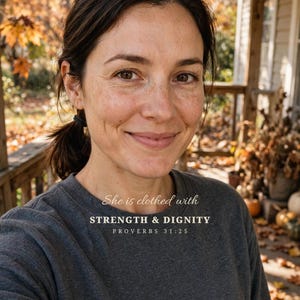 May include: A dark gray long-sleeve crewneck sweatshirt featuring the white printed text: She is clothed with STRENGTH & DIGNITY PROVERBS 31:25. The garment has a simple, solid-colored fabric construction with a standard neckline and long sleeves.
