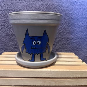 May include: A light gray ceramic plant pot with a matching saucer, featuring a hand-painted illustration of a blue monster with a grumpy expression, pointed ears, and a single visible tooth, centered on the front of the tapered cylindrical vessel.