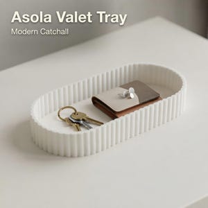 Op de afbeelding: Een ovale witte opbergschaal met een geribbelde buitenkant. In de schaal liggen een kleine beige en bruine portemonnee en een set metalen sleutels aan een goudkleurige ring. De tekst Asola Valet Tray Modern Catchall is zichtbaar.