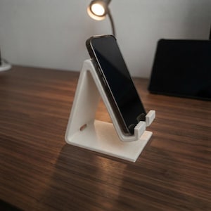 Puede incluir: Un soporte para smartphone blanco en forma de L con un acabado de superficie texturizado y en capas. El soporte cuenta con un pequeño borde saliente en la base para sostener un smartphone negro colocado verticalmente.