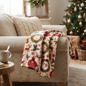 Pode incluir: Uma manta de cor creme com um padrão festivo de renas vermelhas e castanhas, folhas de azevinho verdes, ornamentos vermelhos e bonecos de neve sorridentes com cartolas, drapeada sobre o braço de um sofá estofado em bege claro.
