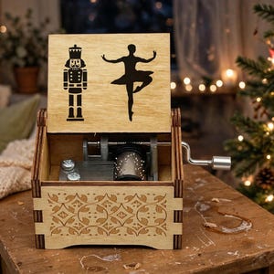 Peut inclure: Une boîte à musique en bois clair avec un couvercle à charnière présentant une silhouette noire de casse-noisette et de ballerine. Le panneau avant affiche un motif floral découpé au laser. Une manivelle en métal argenté est fixée sur le côté.
