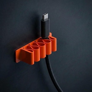 Op de afbeelding: Een feloranje wand-kabelhouder met een reeks herhalende, golfachtige lussen die zijn ontworpen om een enkele zwarte USB-C-oplaadkabel op zijn plek te houden. De houder is bevestigd aan een donker, effen verticaal oppervlak.