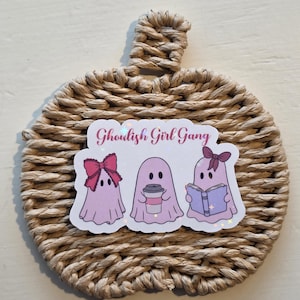 May include: A sticker featuring three light pink ghosts with the text Ghoulish Girl Gang. The first ghost wears a large pink bow, the second holds a coffee cup, and the third wears a small bow while reading a book. The sticker is on a woven pumpkin.