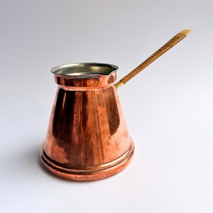 May include: A polished copper coffee pot with a wide base and a narrow neck. The pot features a metallic silver-toned interior rim and a long, slender handle wrapped in a light brown textured material, extending diagonally from the side of the body.