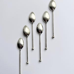 Könnte beinhalten: Ein Set aus fünf silberfarbenen Löffeln, die in einer diagonalen Linie angeordnet sind. Jeder Löffel hat eine ovale Laffe und einen langen, schlanken Stiel mit gedrehtem Metalldesign, der in einer kleinen, glatten Kugel am Ende endet.