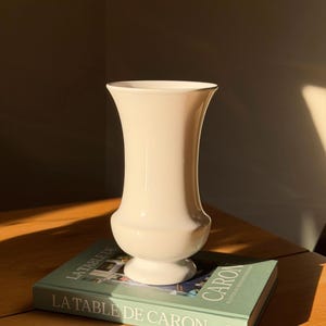 Op de afbeelding: Een gladde, gebroken witte keramische vaas met een uitlopende rand en een afgeronde voet, rustend op een groen hardcover boek getiteld LA TABLE DE CARON, met een fotografische omslag en witte tekst op de rug en de voorzijde van het boek.