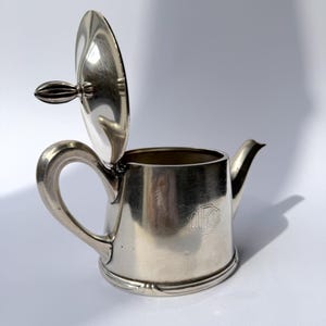 Op de afbeelding: Een zilverkleurige metalen roomkan met een scharnierend deksel, een gebogen handvat en een kleine tuit. De behuizing van de kan is voorzien van een gegraveerd monogram met de letters 'LD' binnen een zeshoekige rand, met een gepolijste afwerking.