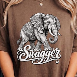 May include: A brown short-sleeve t-shirt featuring a detailed black and white line art illustration of an elephant with intricate patterns on its body. Below the elephant, the word "Swagger" is printed in a bold, white, cursive script font.