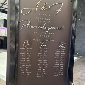 May include: A dark brown wedding seating chart sign with white and gold lettering. The text reads A & F, 09.07.26, Tyne Bank Brewery, Please take your seat, Sweetheart Table, Bride, Groom, and lists guest names under tables One, Two, and Three.