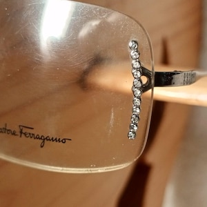 Może przedstawiać: Okulary przeciwsłoneczne bez oprawek z przezroczystymi soczewkami, z czarnym nadrukiem logo Salvatore Ferragamo na prawej soczewce. Pionowy rząd małych, przezroczystych, okrągłych cyrkonii jest osadzony wzdłuż zewnętrznej krawędzi przy zauszniku.