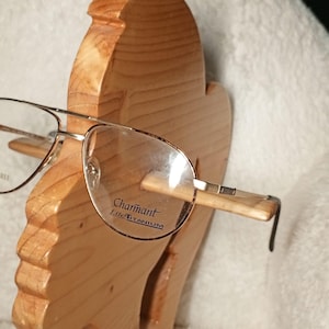 Puede incluir: Gafas de estilo aviador con monturas metálicas finas. Los cristales presentan el texto impreso Charmant Lite Titanium y Allergy Free. Las gafas se muestran sobre un soporte de madera tallada con forma de perfil humano.