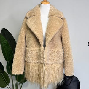 May include: A beige coat with a textured, curly fleece upper section and a long, shaggy fringe hem. The coat features a wide notched collar, long sleeves, and a straight-cut silhouette, displayed on a white mannequin torso stand.