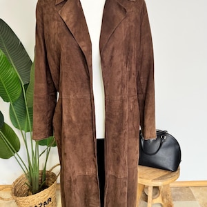 Peut inclure: Un long manteau en daim marron foncé doté d'un col à revers, de manches longues et d'une coupe droite ouverte sur le devant. Le vêtement présente des coutures horizontales visibles au niveau de la taille et du bas, créant une silhouette structurée.