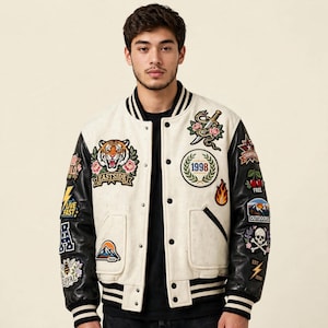 Könnte beinhalten: Eine cremefarbene Collegejacke mit schwarzen Lederärmeln, die einen gerippten Kragen und Bündchen mit schwarzen und weißen Streifen aufweist. Die Jacke ist mit verschiedenen gestickten Aufnähern verziert, darunter ein Tiger, eine Schlange mit Dolch und der Text 1998.