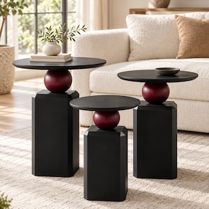 Puede incluir: Un conjunto de tres mesas auxiliares, cada una con un tablero circular negro apoyado sobre una pieza decorativa esférica de color rojo oscuro y una base de pedestal octogonal negra. Las mesas varían en altura, creando una disposición escalonada.
