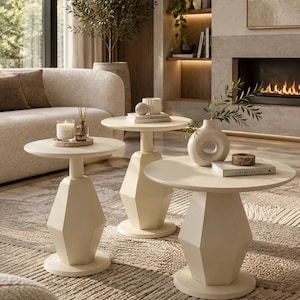 Può includere: Un set di tre tavolini color bianco sporco, ciascuno caratterizzato da un piano circolare sostenuto da una base a piedistallo geometrica e sfaccettata. I tavoli sono disposti ad altezze variabili e presentano una finitura liscia e opaca.