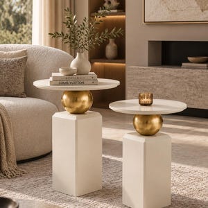 May include: Two side tables featuring off-white square pedestals, gold spherical accents, and circular off-white tabletops. The taller table holds a white vase with greenery and two books titled The Kinfolk Home and Louis Vuitton.