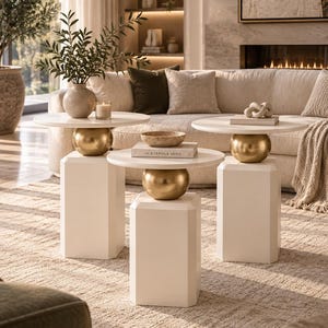 May include: A set of three side tables, each featuring a smooth white circular top, a central gold-toned spherical accent, and a white hexagonal pedestal base. The tables are arranged in a cluster, with one table holding a vase and candle.