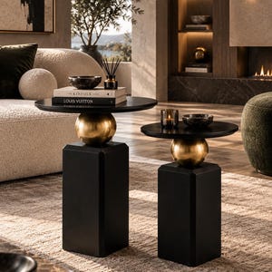 May include: Two side tables featuring circular black tabletops supported by a central gold-toned sphere and a solid black rectangular base. The tables are of varying heights and display a smooth, matte finish on the black components.