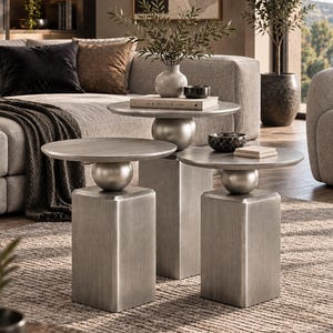May include: A set of three nesting side tables with a brushed silver metallic finish. Each table features a circular top supported by a central spherical accent piece resting on a rectangular pedestal base. The tables are arranged in a tiered formation.