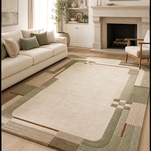 May include: A rectangular area rug featuring a large cream-colored center panel surrounded by a geometric border in shades of sage green, tan, and brown. The border includes textured rectangular blocks, linear patterns, and rounded corner accents.