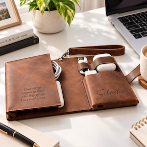 Op de afbeelding: Een bruine, lederachtige tech-organizer met polsband, voorzien van de gegraveerde tekst "Good things happen to those who keep going. I'm proud of you." en "Sebastian." Het bevat vakjes voor een witte oplaadkabel, een stroomadapter en witte oordopjes.
