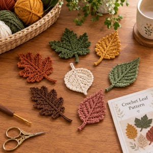 May include: A collection of seven crocheted leaves in various shapes and colors, including green, yellow, brown, cream, and pink. A crochet hook, a pair of scissors, a basket of yarn, and a booklet titled Crochet Leaf Pattern are also visible.
