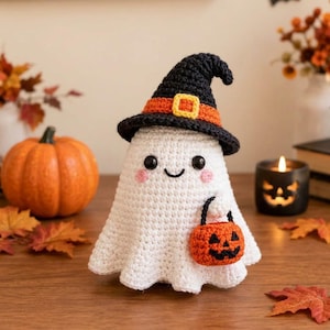 May include: A white crocheted ghost figurine wearing a black witch hat with an orange band and yellow buckle. The ghost has black bead eyes, pink cheeks, and a stitched smile, and is holding a small orange crocheted pumpkin treat bucket.