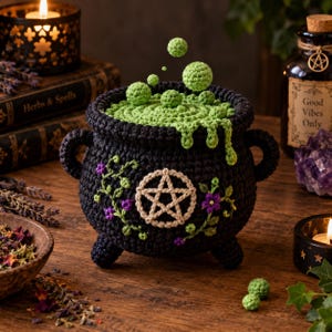 Op de afbeelding: Een zwarte gehaakte ketel met een beige pentagram-embleem aan de voorzijde, versierd met groene ranken en paarse bloemen. De ketel is gevuld met een getextureerde groene vloeistof en zwevende groene bollen.