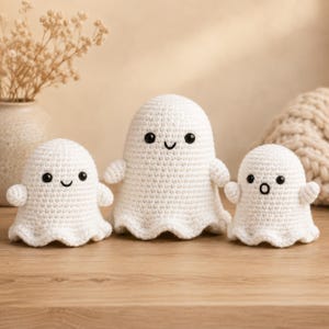 Puede incluir: Tres figuras de fantasmas tejidas a ganchillo en color blanco con cuerpos redondeados y bordes inferiores ondulados. Cada fantasma tiene dos pequeños ojos negros circulares y una boca bordada. El fantasma central sonríe y los otros tienen expresiones.