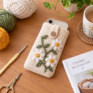 May include: A cream-coloured crochet phone pouch featuring a floral design with white daisy petals and yellow centres, green stems, and leaves. The pouch has a button closure at the top and contains a smartphone. The texture is thick and yarn-based.