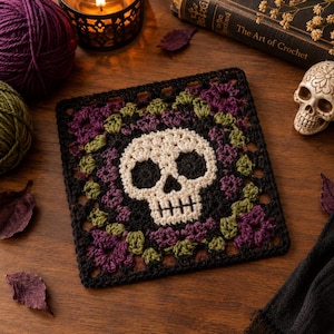 May include: A square crochet granny square featuring a central off-white skull motif with black eye sockets and nose. The skull is surrounded by a border of dark purple and olive green floral-like crochet patterns, all framed by a black yarn border.