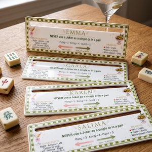 Pode incluir: Quatro suportes retangulares brancos para peças de Mahjong com bordas xadrez verdes, personalizados com os nomes Emma, Carla, Karen e Salima. Os suportes apresentam instruções de jogo impressas, incluindo regras de Charleston e códigos de cor.