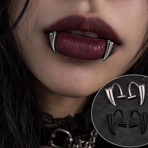 Può includere: Un set di piercing per labbra a forma di zanna in tonalità argento e nero. Il gioiello presenta un design curvo con un'estremità appuntita, che ricorda un dente di vampiro. La versione argentata è mostrata indossata sul labbro inferiore.
