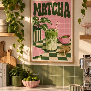 May include: A framed wall art print featuring the word MATCHA in large dark green letters. The illustration depicts a glass of iced matcha latte, a potted plant, a small pink bowl of matcha powder, and a bamboo whisk on a green and white checkered surface.