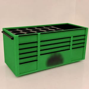 May include: A bright green desk organizer shaped like a professional rolling tool chest. It features multiple horizontal drawer-style indentations with black trim, a side handle, and a top surface with circular holes for holding pens or tools.
