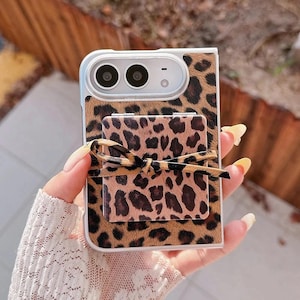 May include: A rectangular phone case featuring a brown and black leopard print pattern. The case includes a matching square-shaped pop-out grip on the back, which is adorned with a small, tied leopard print ribbon centred on its surface.
