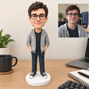 Op de afbeelding: Een op maat gemaakt bobblehead-figuurtje van een persoon met bruin haar en een bril met zwart montuur, gekleed in een grijs vest met capuchon over een zwart t-shirt, een donkerblauwe spijkerbroek en zwart-witte sneakers op een witte voet.