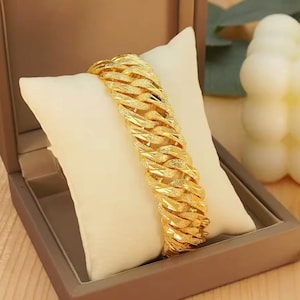 Puede incluir: Una pulsera de cadena tipo barbada en tono dorado con eslabones gruesos entrelazados y un acabado texturizado y brillante. La pulsera se muestra en diagonal sobre un cojín cuadrado de tela color blanco roto, destacando su brillo metálico.
