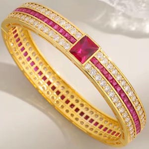 May include: A gold-toned hinged bangle bracelet featuring a central rectangular red gemstone flanked by two rows of clear white stones and two rows of square-cut red stones. The design includes a repeating pattern of red and white stones throughout.