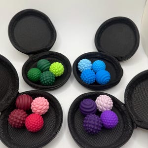 May include: Four round black zippered cases, each containing a set of four small, textured silicone sensory balls. The balls come in various colors, including shades of green, blue, pink, red, and purple, featuring bumpy and ridged surfaces.