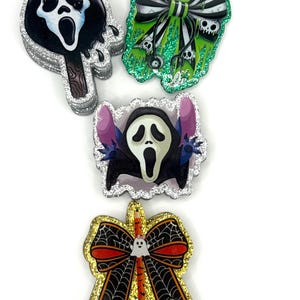 Könnte beinhalten: Ein Set aus vier dekorativen Acryl-Ansteckern mit Glitzerrändern. Die Motive umfassen ein schwarz-weißes Geistermasken-Eis am Stiel, eine grüne Schleife mit Totenköpfen, eine Geistermasken-Figur mit lila Ohren und eine orangefarbene Schleife mit Spinnennetz.