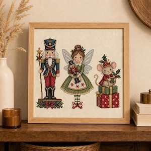 May include: A framed cross-stitch embroidery featuring a nutcracker in a blue and red uniform, a fairy in a green dress holding a small nutcracker, and a mouse wearing a red cape sitting on two stacked gift boxes with holly accents.