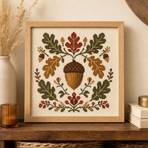 May include: A square cross-stitch embroidery in a light wood frame featuring a central brown acorn surrounded by symmetrical oak leaves in shades of green, gold, and rust, along with small floral accents on a cream-colored fabric background.
